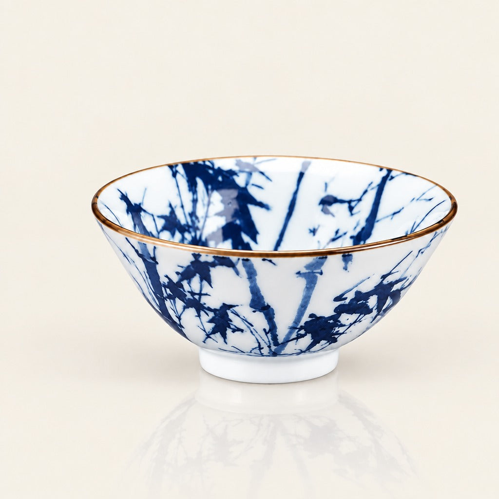 A white ceramic bowl decorated with a blue bamboo pattern and a thin gold rim, shown on a clean off-white background with a soft reflection below.