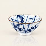 A white ceramic bowl decorated with a blue bamboo pattern and a thin gold rim, shown on a clean off-white background with a soft reflection below.