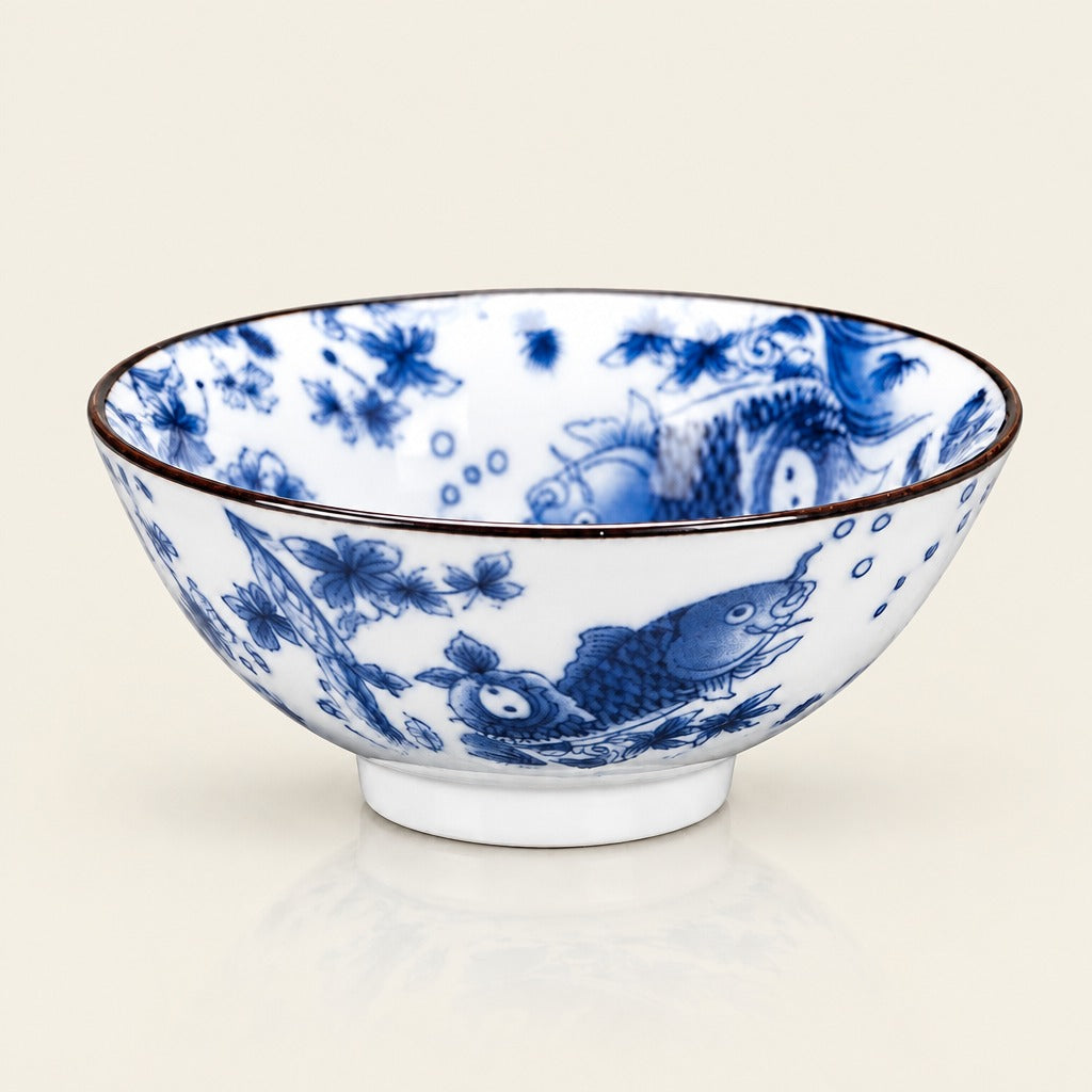 A white ceramic bowl decorated with blue floral and fish motifs, featuring a dark brown rim and a small white pedestal base, set against a light cream background.