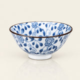 A white ceramic bowl decorated with a blue floral and leaf pattern, featuring a thin dark brown rim and a small white footed base, set against a warm off-white background.