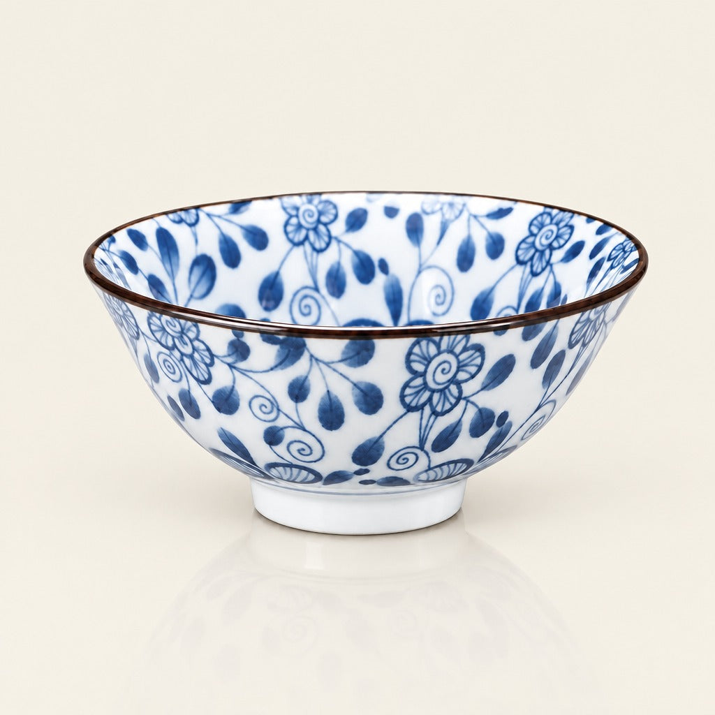 A white ceramic bowl decorated with a blue floral and leaf pattern, featuring a thin dark brown rim and a small white footed base, set against a warm off-white background.
