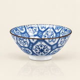 A ceramic bowl with a blue and white floral geometric pattern, featuring a thin dark rim and a small white footed base, set against a solid cream background.