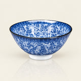 A blue and white floral pattern ceramic bowl with a black rim, displayed on a clean, light beige reflective surface.