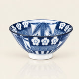 A blue and white patterned ceramic bowl with a small white footed base, photographed on a clean light beige background with a soft reflection below.