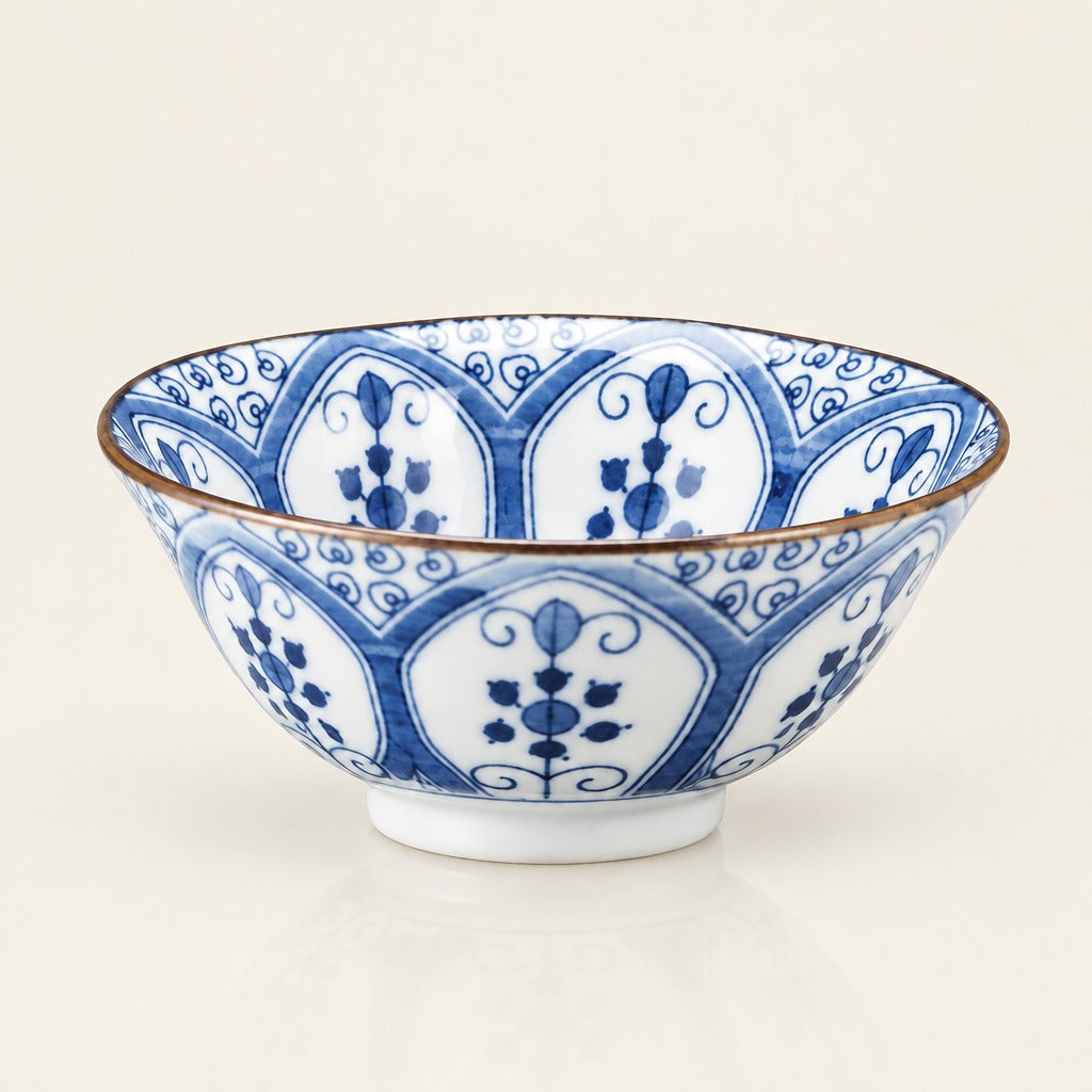A white ceramic bowl with a blue floral and scroll pattern, featuring a thin brown rim and a small white footed base, photographed on a clean off-white background.