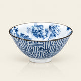 A white ceramic bowl decorated with blue hand-painted landscape scenes and a blue geometric pattern on the exterior, finished with a thin gold rim, set against a light cream background.
