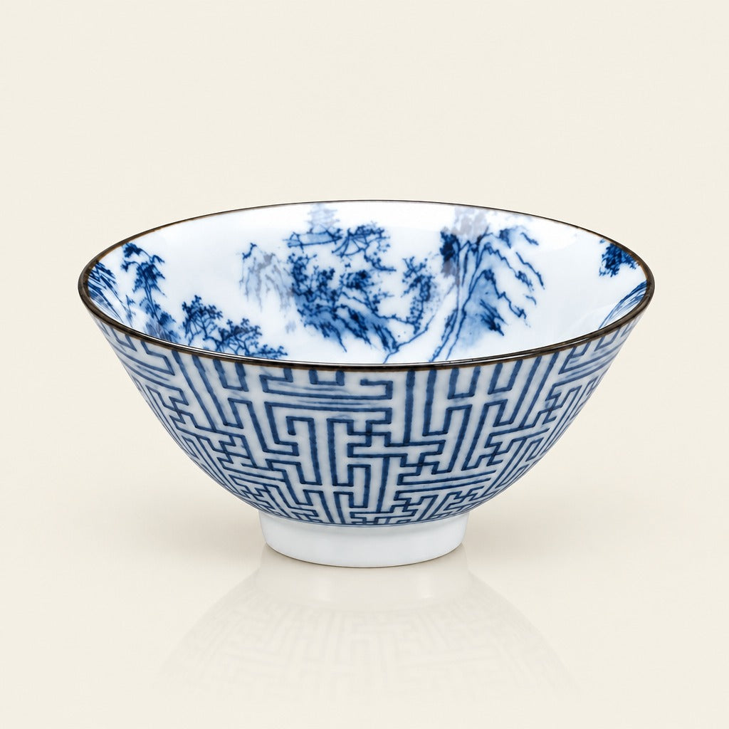 A white ceramic bowl decorated with blue hand-painted landscape scenes and a blue geometric pattern on the exterior, finished with a thin gold rim, set against a light cream background.