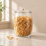 A large clear glass jar with a matching glass lid and a small gold-toned knob, filled with golden cereal pieces, sitting on a light marble countertop in a softly lit room.