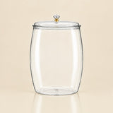 A clear glass canister jar with a matching glass lid and a small gold-toned knob, shown on a warm beige background.