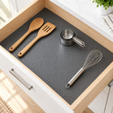 A dark gray textured kitchen drawer mat laid inside an open wooden drawer, styled with wooden spoons, metal measuring cups, and a whisk.