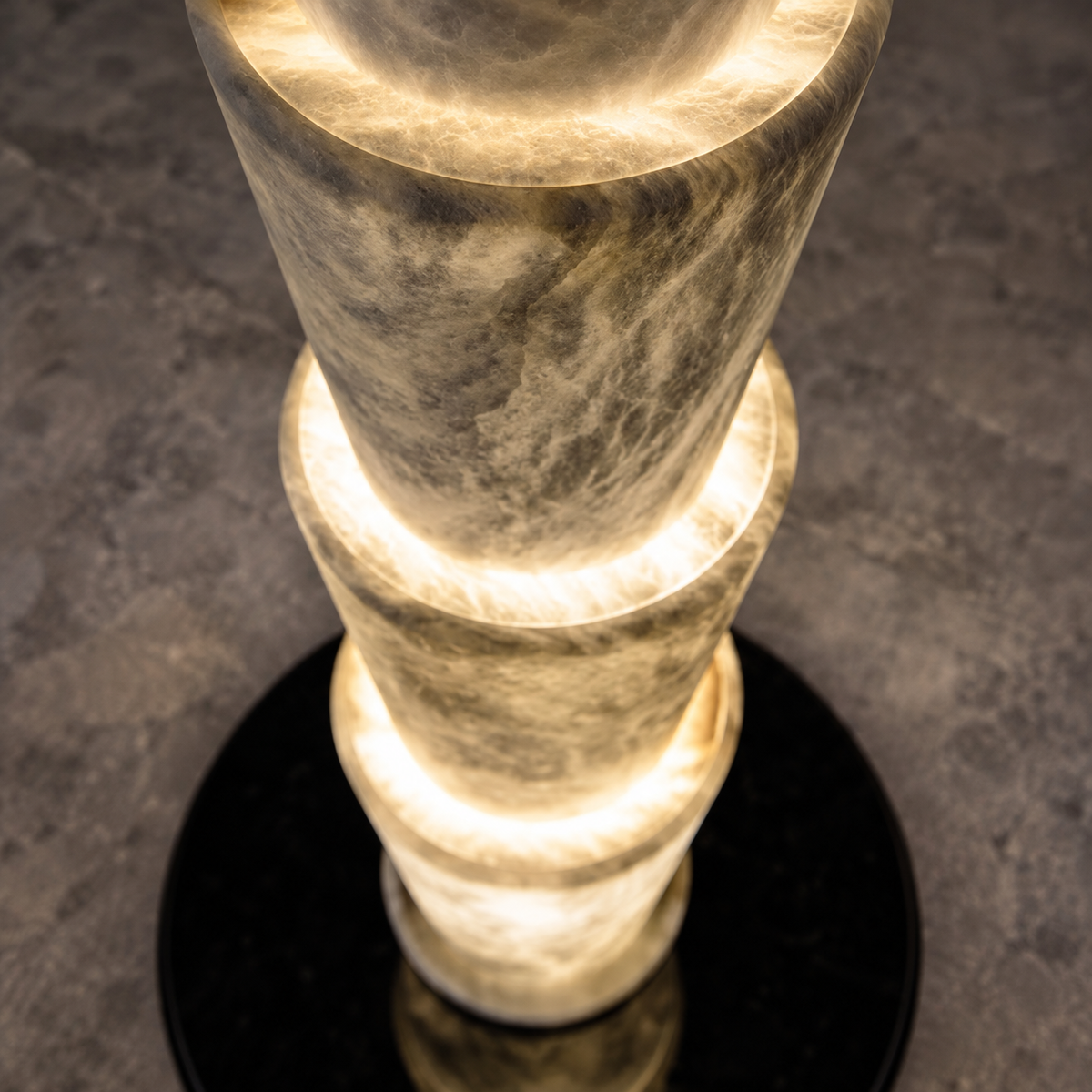 A modern sculptural floor lamp made of stacked, glowing stone or acrylic cylinders on a round black base, photographed from above against a dark textured floor.