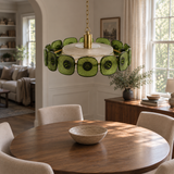 A modern circular pendant chandelier with green textured glass discs and a brass center, hanging over a round wooden dining table in a bright, modern dining room.