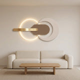 Modern wall-mounted light fixture with a glowing circular ring and a horizontal brushed-gold bar, mounted above a cream sofa in a minimalist living room.