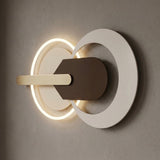 Modern wall-mounted light fixture with two overlapping circular rings, one glowing warm white, a white outer ring, a dark brown central panel, and a horizontal cream bar against a textured gray wall.