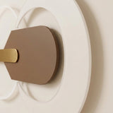 Wall light close-up showing a taupe central plate, gold-tone accent, and layered white circular forms against a cream wall.