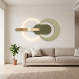 Modern wall-mounted light fixture with a glowing circular ring, a horizontal brushed gold bar, and overlapping matte green circular and leaf-shaped elements, shown above a beige sofa in a bright living room.