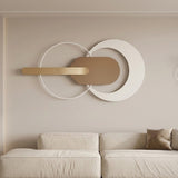 Modern wall-mounted abstract decor piece with interlocking white and tan geometric shapes and a horizontal gold bar, displayed above a light beige sofa in a minimalist living room.
