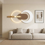 Modern wall-mounted light fixture with a glowing circular ring, a white crescent shape, and a horizontal brushed-gold bar, displayed above a light beige sofa in a minimalist living room.