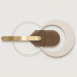 A modern wall-mounted light fixture with overlapping circular and curved geometric elements in beige, brown, and brushed gold metal, shown against a clean off-white background.