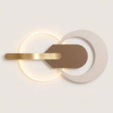 Modern wall sconce with a glowing circular light, a horizontal brushed gold bar, and overlapping matte white and taupe geometric shapes on a light neutral background.