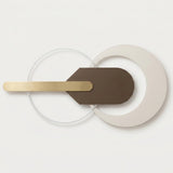 Modern wall-mounted light fixture with a matte brown geometric central plate, a brushed gold horizontal bar, and overlapping translucent and off-white circular rings on a clean white background.