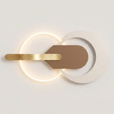 Modern wall sconce with a glowing circular LED light, a brushed gold horizontal bar, and a matte taupe geometric shield overlay on a white circular plate, shown against a neutral off-white background.