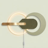 Modern wall-mounted light fixture with a glowing circular ring, a large matte off-white circular cutout, a dark olive green geometric center, and a horizontal brushed brass bar, shown against a pale beige wall.