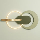 Modern wall-mounted light fixture with a glowing circular ring, a large matte off-white crescent shape, a dark olive green central shield, and a horizontal brushed brass bar against a light neutral wall.