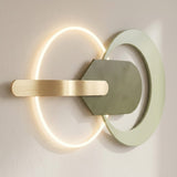 Wall light with a glowing warm-white ring, sage-green circular frame and central plate, and horizontal gold-tone bar against a beige wall.