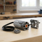 A grey building block motor component with coiled black cable sits beside a gear assembly on a light wooden workbench.