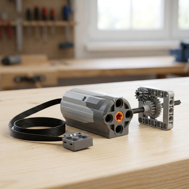 A grey building block motor component with coiled black cable sits beside a gear assembly on a light wooden workbench.