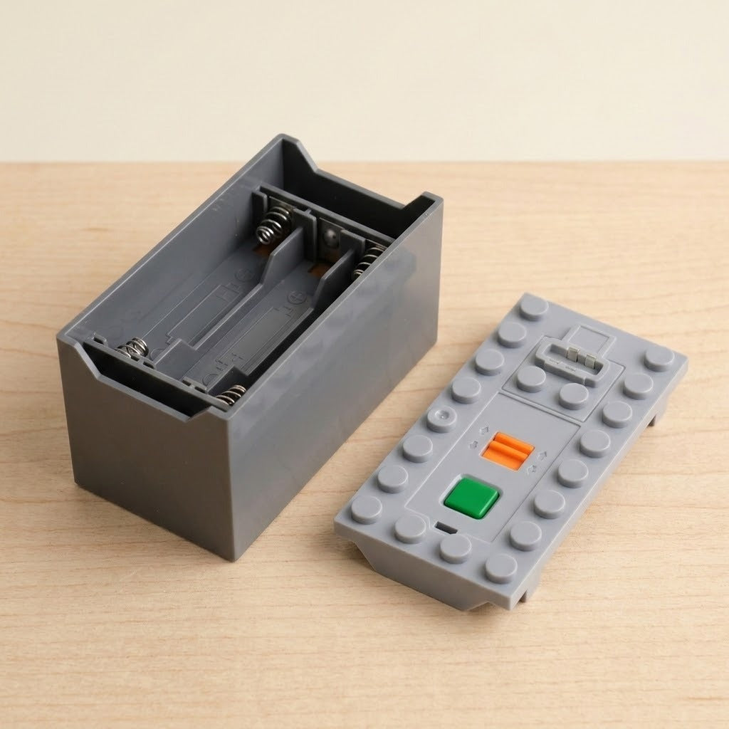 A gray plastic building block battery component sits open beside its studded lid with green and orange controls on a wood tabletop.