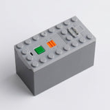 A gray electronic module or control box with a top stud base, featuring a green button, an orange switch, and a small gray connector on a light gray background.