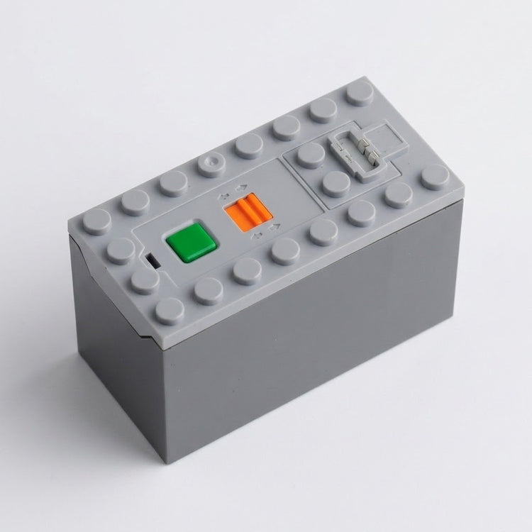 A gray electronic module or control box with a top stud base, featuring a green button, an orange switch, and a small gray connector on a light gray background.
