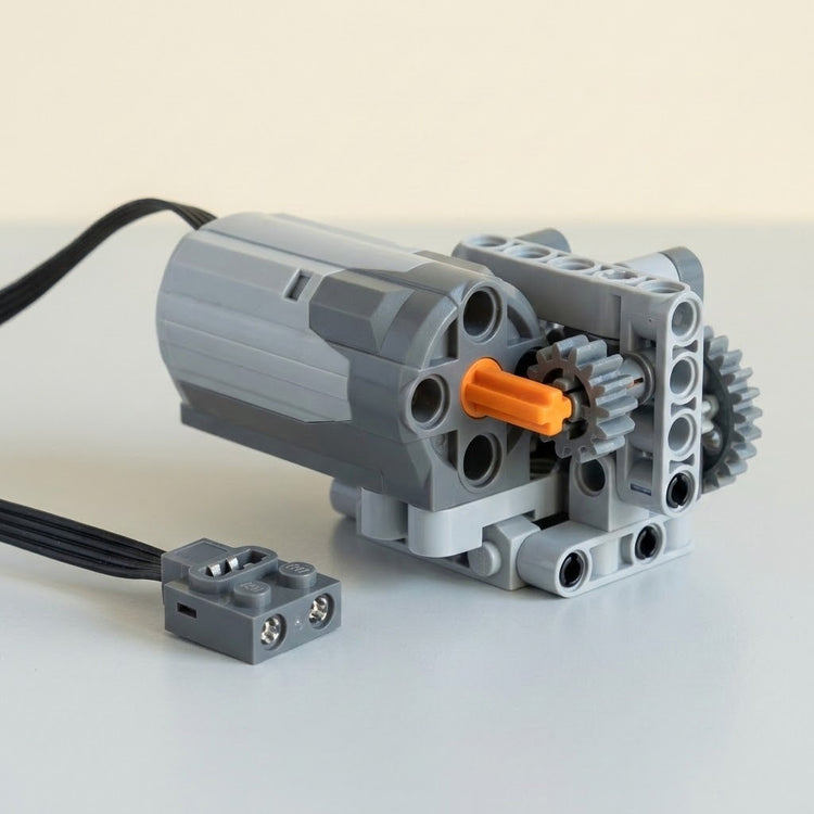 A grey building block motor component connects to a demonstration gear assembly with an orange axle on a pale surface.