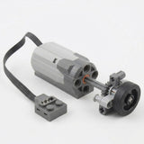 A grey building block motor component with black cable connects to a demonstration wheel and gear assembly against a white background.