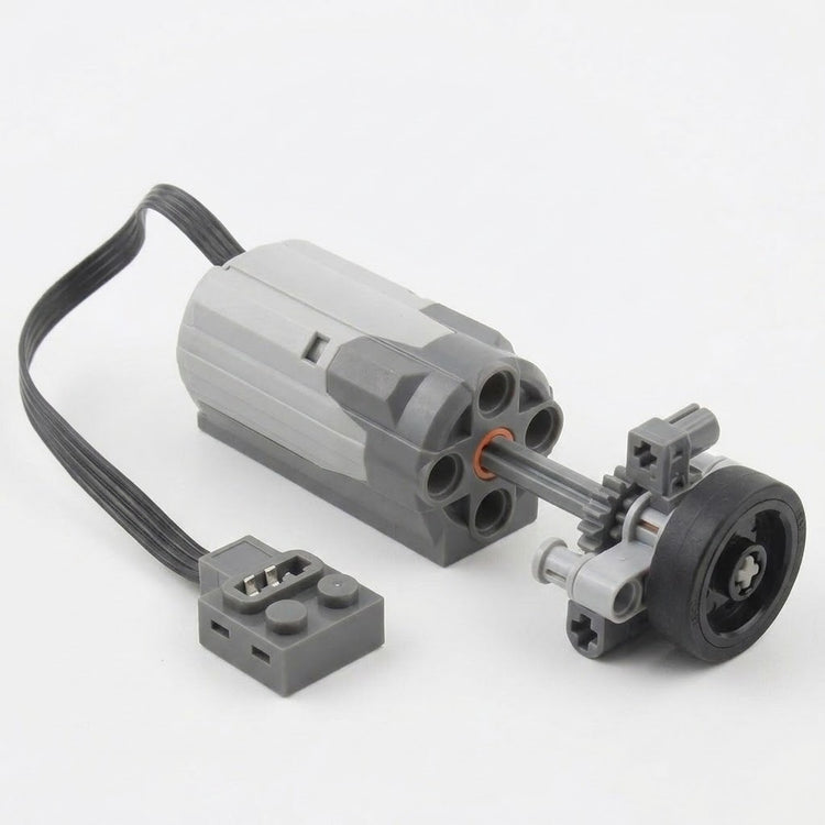 A grey building block motor component with black cable connects to a demonstration wheel and gear assembly against a white background.