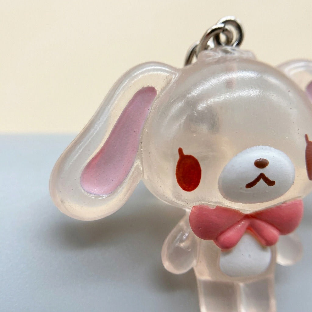 A close-up of a transparent acrylic bunny keychain charm with pink inner ears, a pink bow, and a silver metal ring at the top, set against a split beige and gray background.