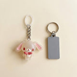 A cute acrylic keychain featuring a small white bunny with pink inner ears and a pink bow tie, shown next to a blank gray rectangular tag on a light beige background.