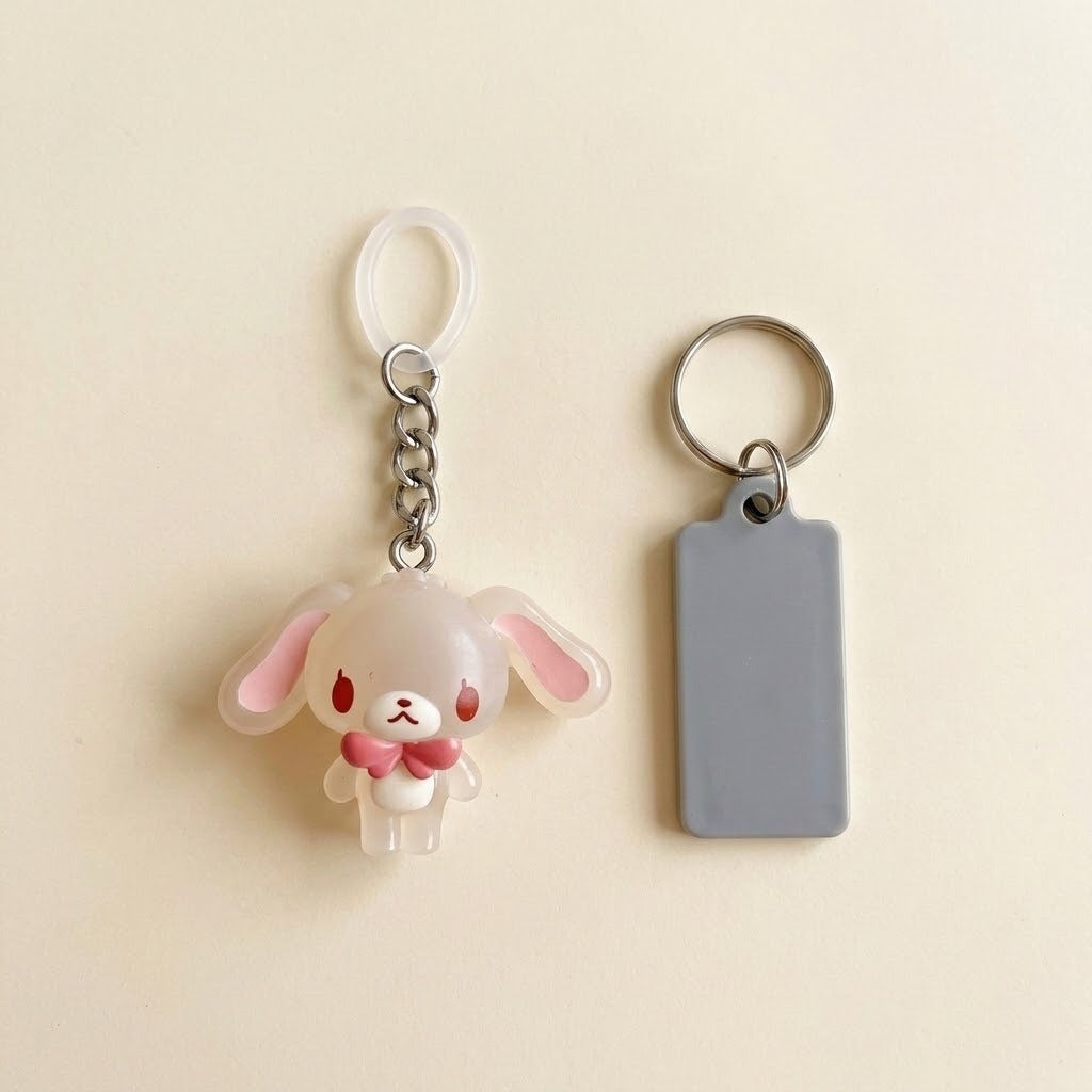 A cute acrylic keychain featuring a small white bunny with pink inner ears and a pink bow tie, shown next to a blank gray rectangular tag on a light beige background.