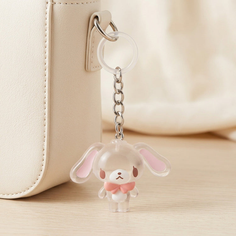 A cute clear acrylic bunny keychain charm with pink inner ears and a pink bow tie, hanging from a silver chain next to a cream-colored handbag.