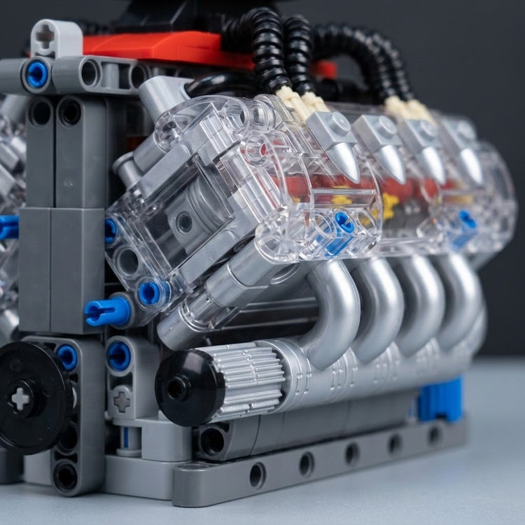 Close-up of a plastic V8 engine model kit showing transparent cylinder housings and curved silver exhaust pipes on a dark background.