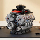 A V8 engine model kit with a silver belt and pulleys, sitting on a dark work mat against a plain light wall.