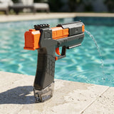 Black and orange water squirt gun stands on a sunlit poolside ledge, releasing an arcing stream toward the right.