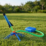 Rocket launcher toy on grass with an angled blue foam rocket, black fins, blue stand and foot pump connected by a green hose.