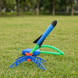 Rocket launcher toy on grass with a blue foam rocket tilted right, green launch tube, orange joint, blue stand and foot pump.