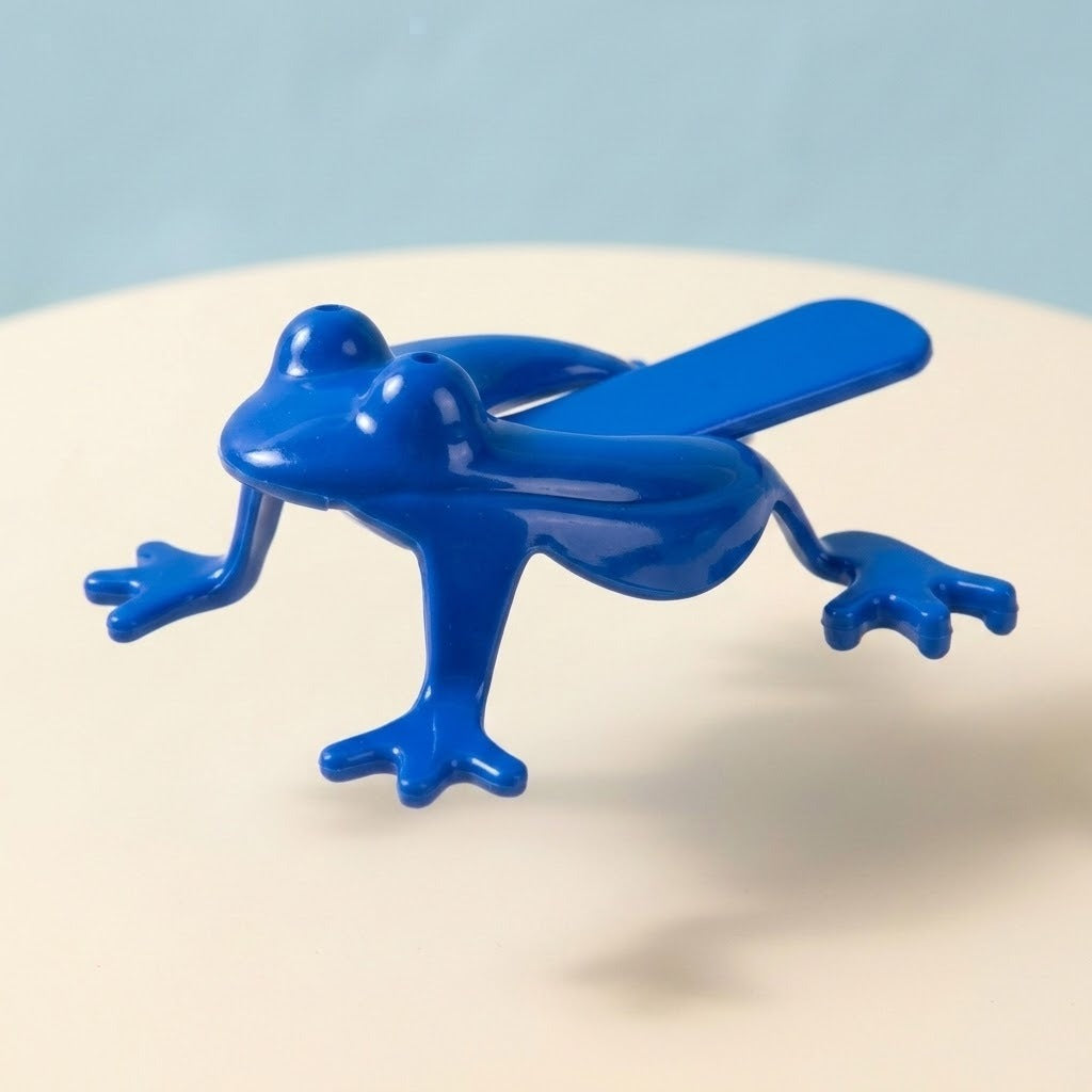 A glossy blue plastic jumping frog toy appears above a cream surface, showing raised eyes, splayed legs and a rear pressing tab.