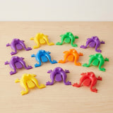 Colorful plastic jumping frog toys with rear pressing tabs sit on a light wooden tabletop against a cream wall.