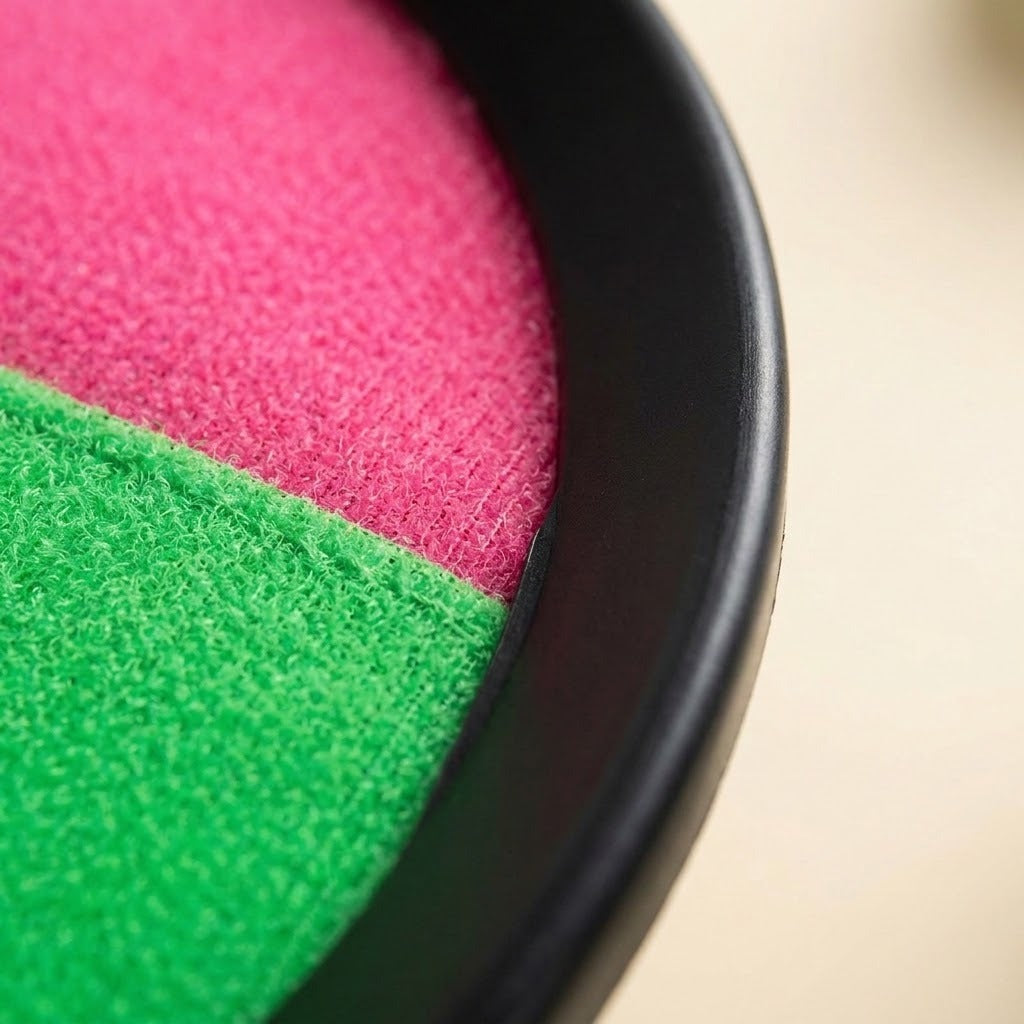 A catch game set paddle close-up shows pink and green textured cloth inside a black plastic rim against a cream background.