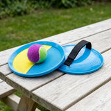 A catch game set with blue paddles, a black hand strap and a purple-and-green ball rests on a weathered picnic table.